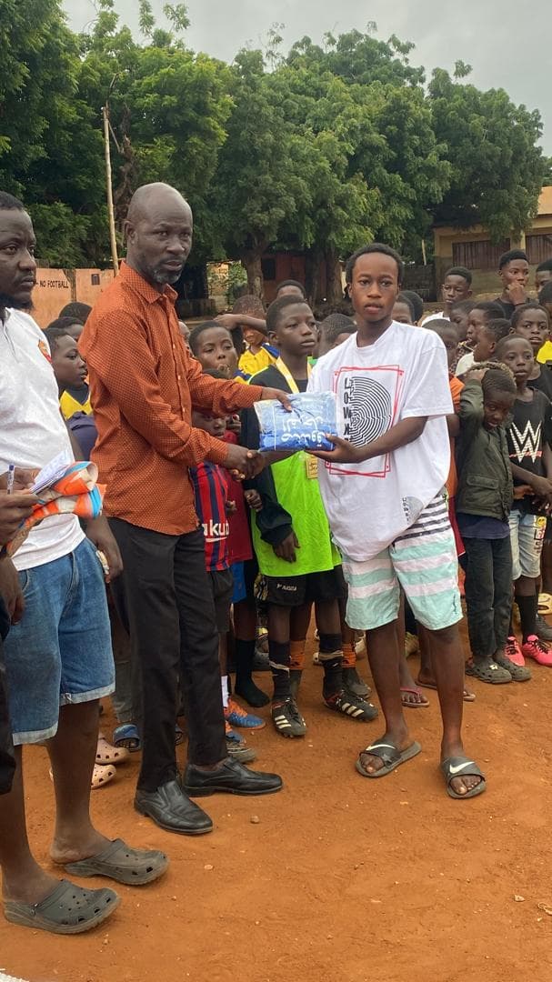 A coach in an orange shirt hands a wrapped gift to a teenage player as kids watch